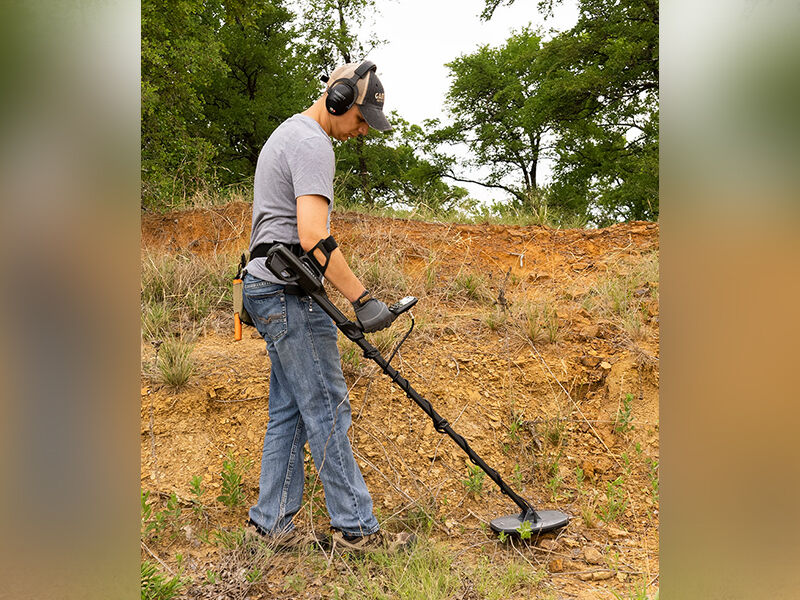 A man is walking along a hillside with exposed red soil. There are grasses and trees in the background. He is wearing a baseball had and headphones. He carries a metal detector and is sweeping the surface of the ground with the flat disk that detects metal. (image courtesy Garrett)